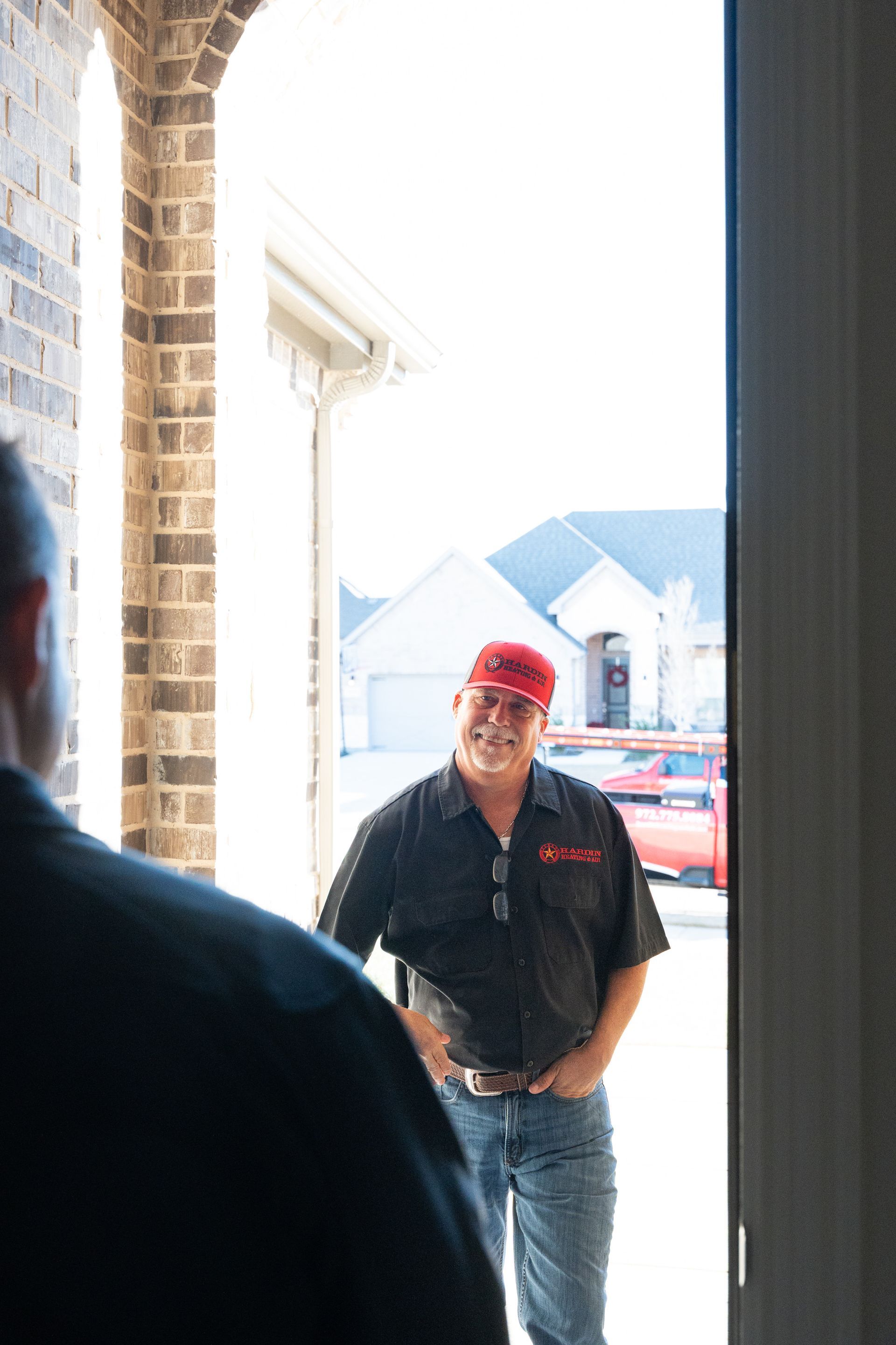 Man in red cap and uniform smiles at a person through an open doorway, sunny outdoor setting.