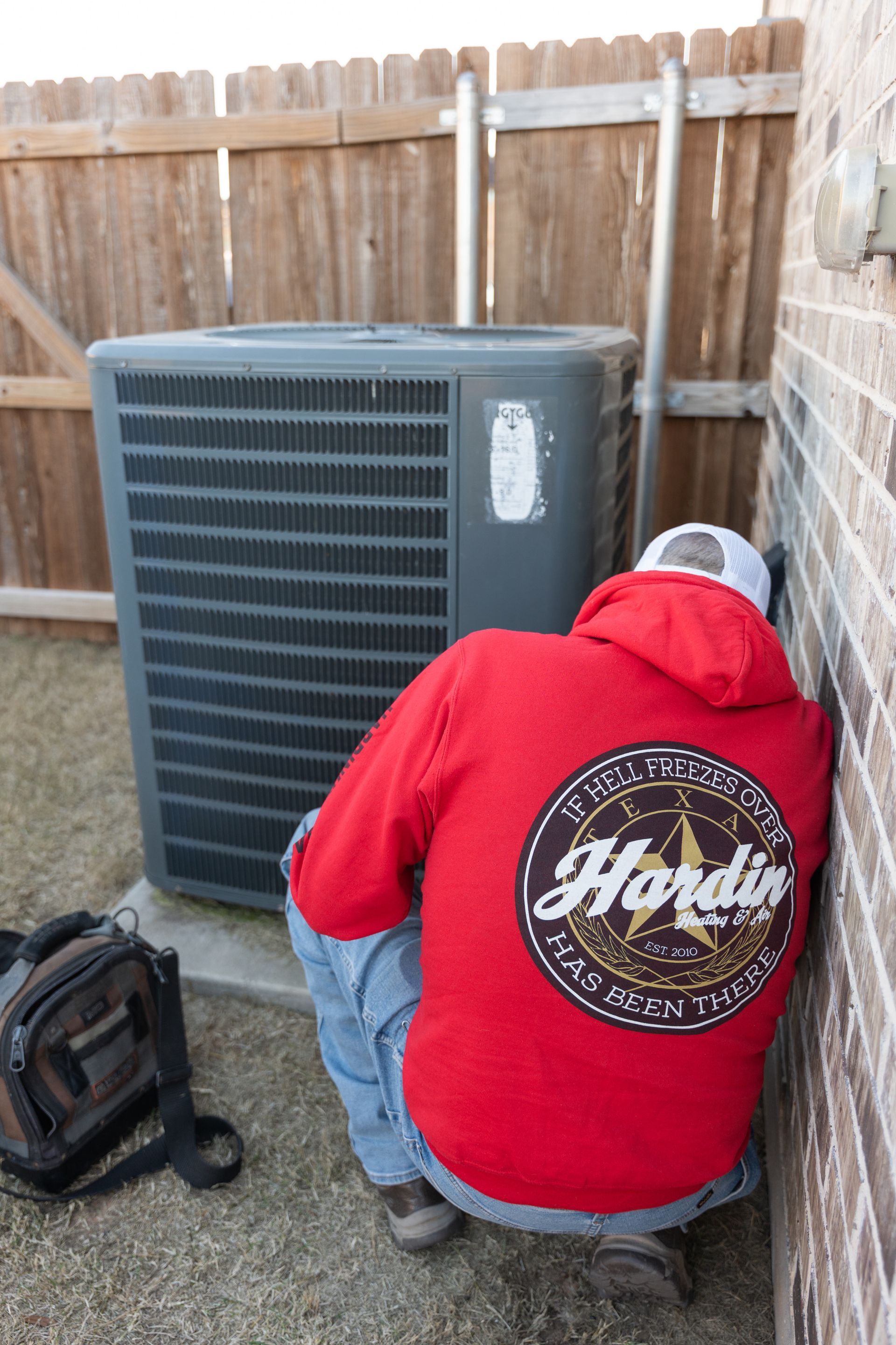 Person in red hoodie inspects an air conditioning unit outside, next to a brick wall and wooden fence.