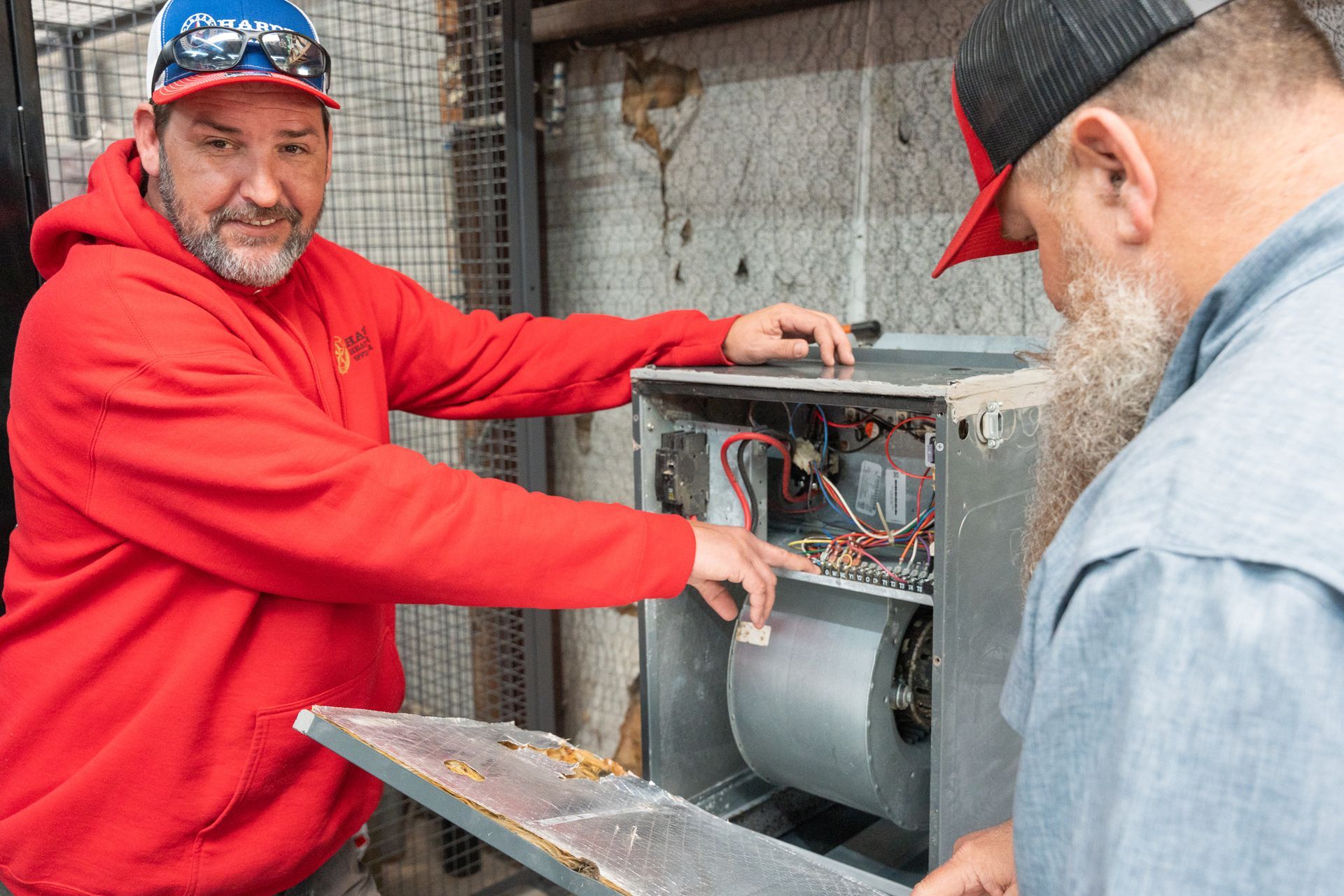 Two Hardin Heating & Air Technician examining an opened HVAC unit; one in red hoodie pointing, other in blue shirt.