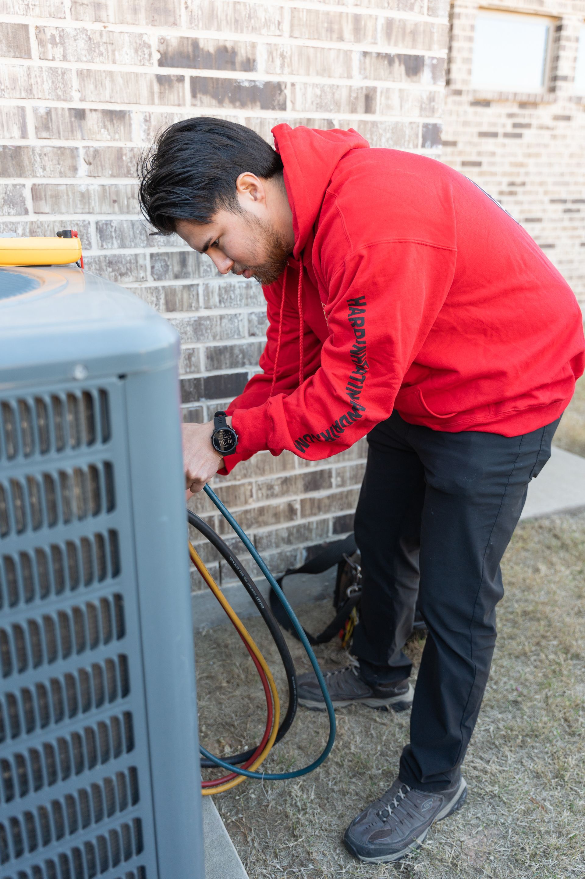 Hardin Heating & Air Technician connects hoses to an AC unit outside a brick building. He wears a red hoodie.
