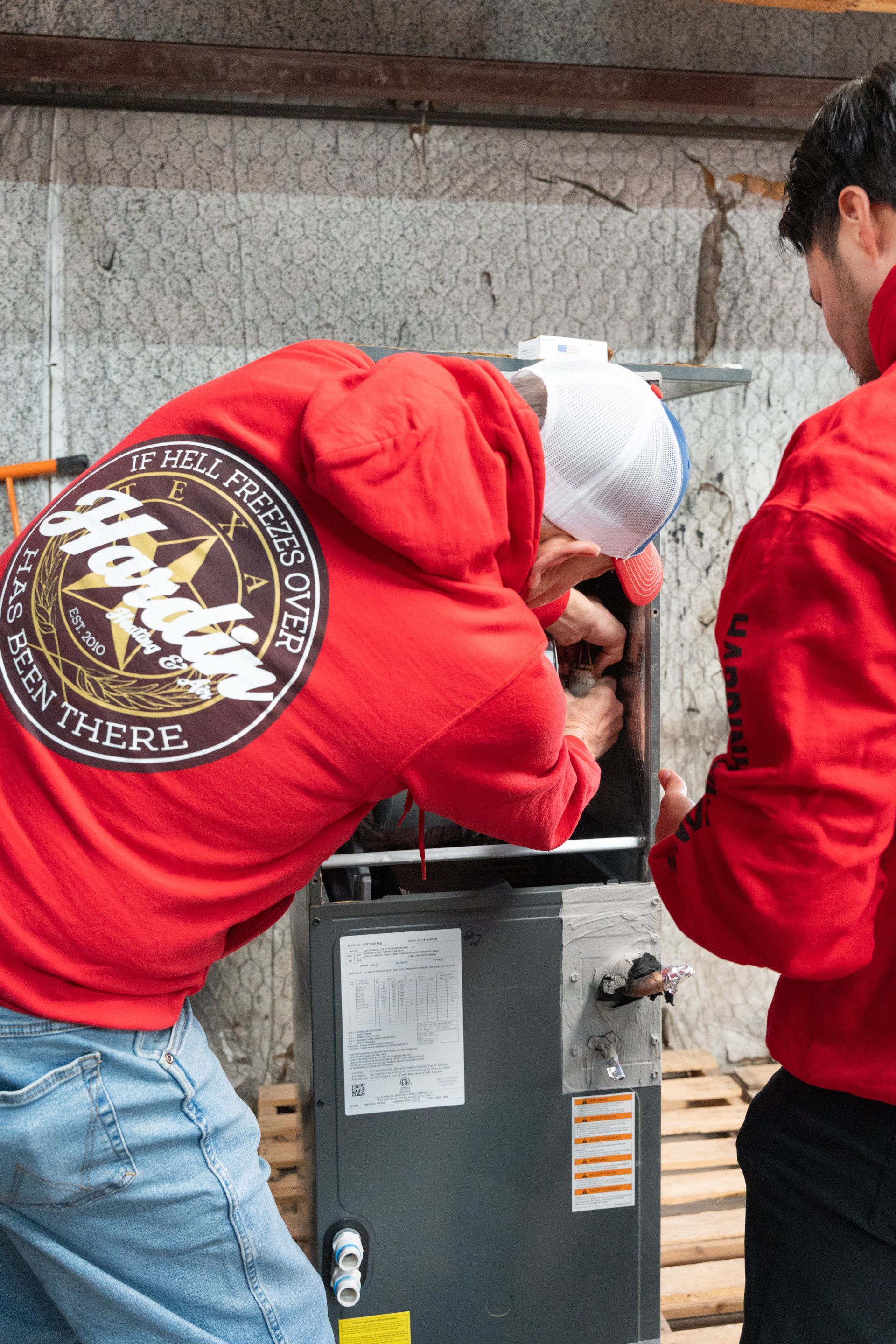 Two Hardin Heating & Air Technician in red hoodies working on HVAC equipment outside.
