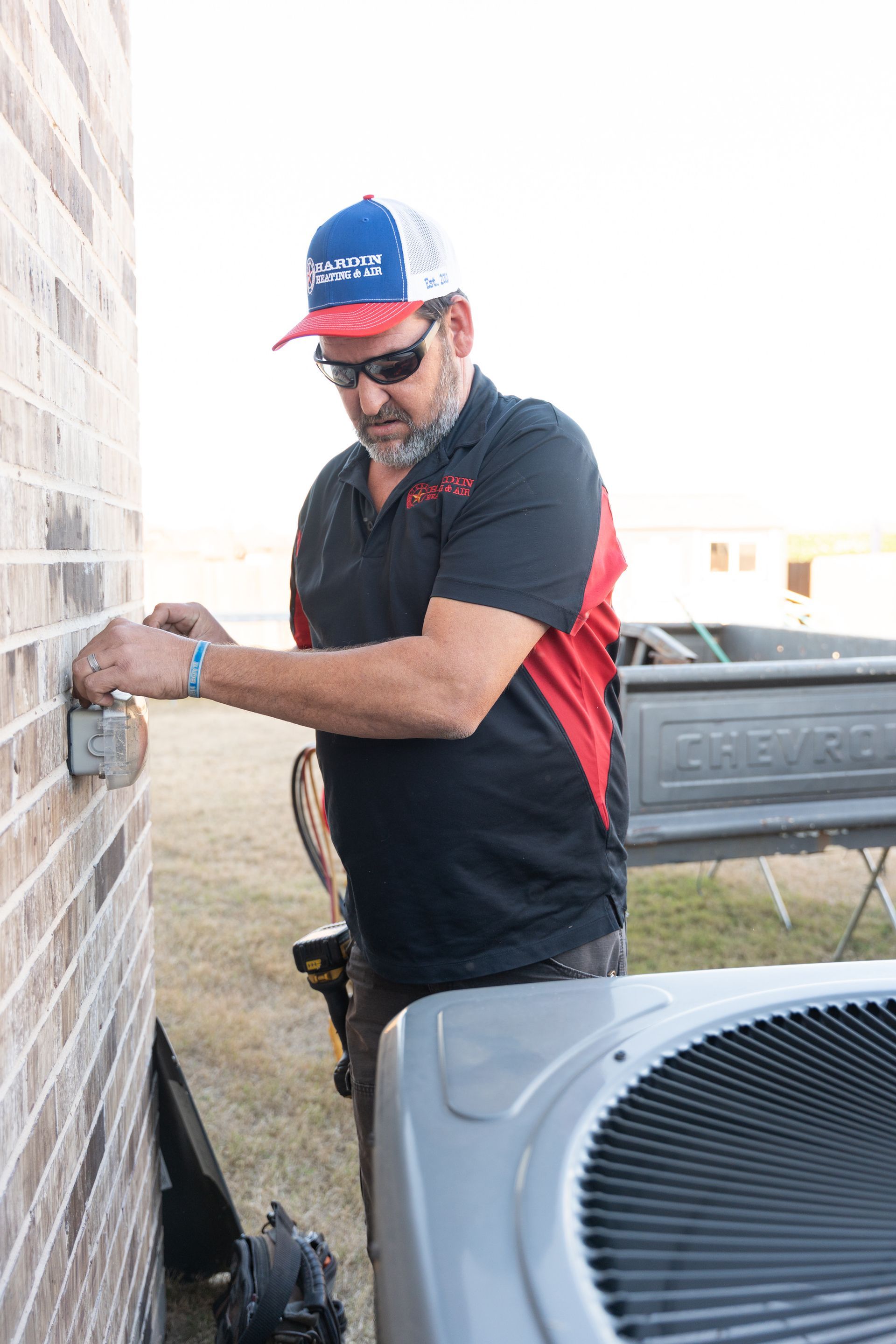 Hardin Heating & Air Technician installing electrical box on brick building near air conditioning unit.