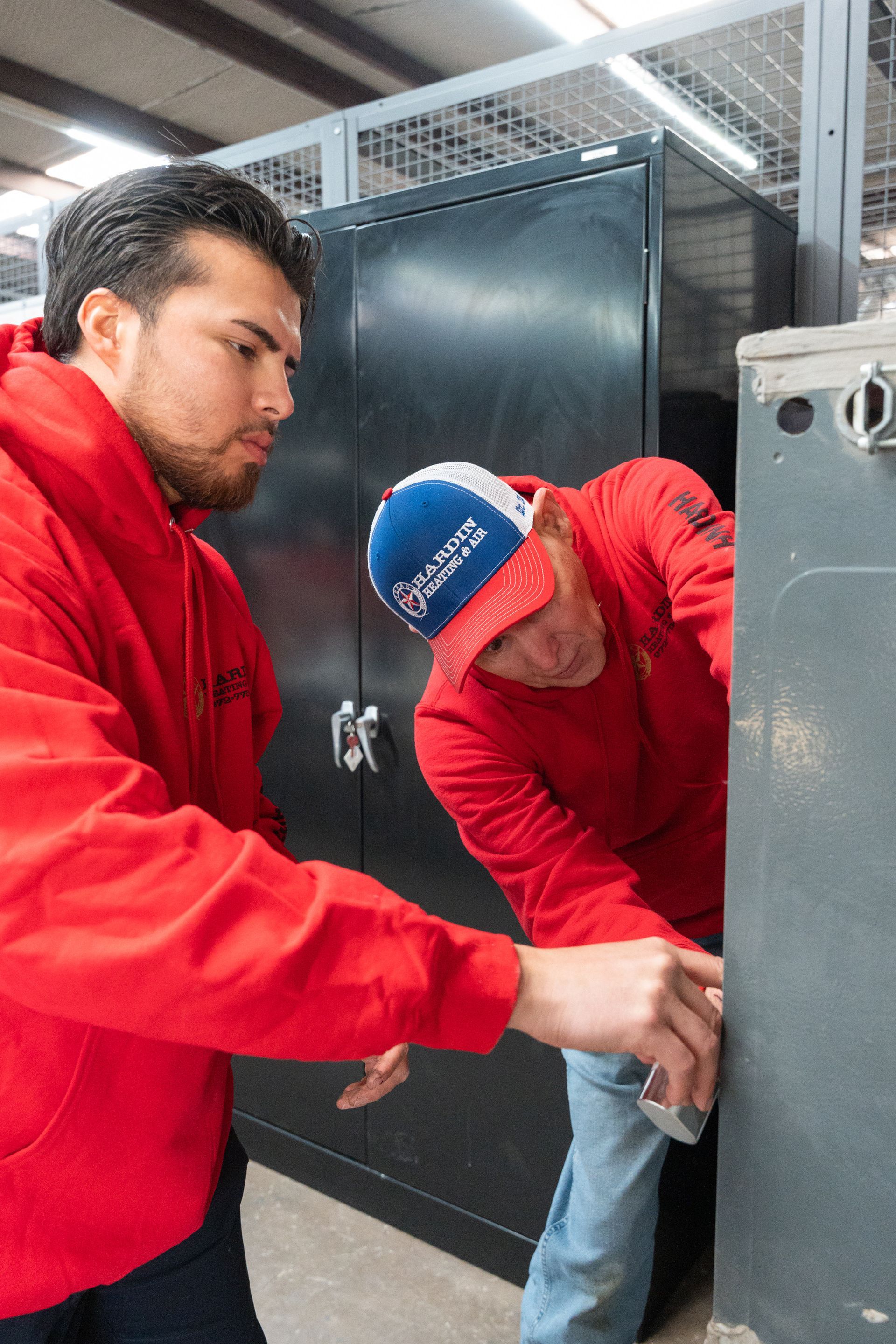 Two Hardin Heating & Air Technician in red hoodies inspect equipment in a workshop, one wearing a hat.