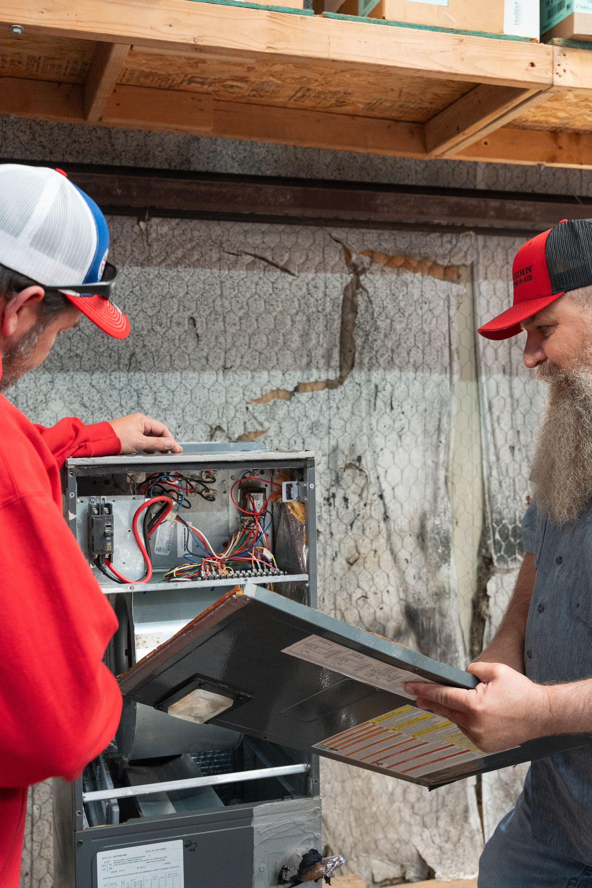 Two Hardin Heating & Air Technician working on electrical panel outdoors; one holds a cover, the other points inside.