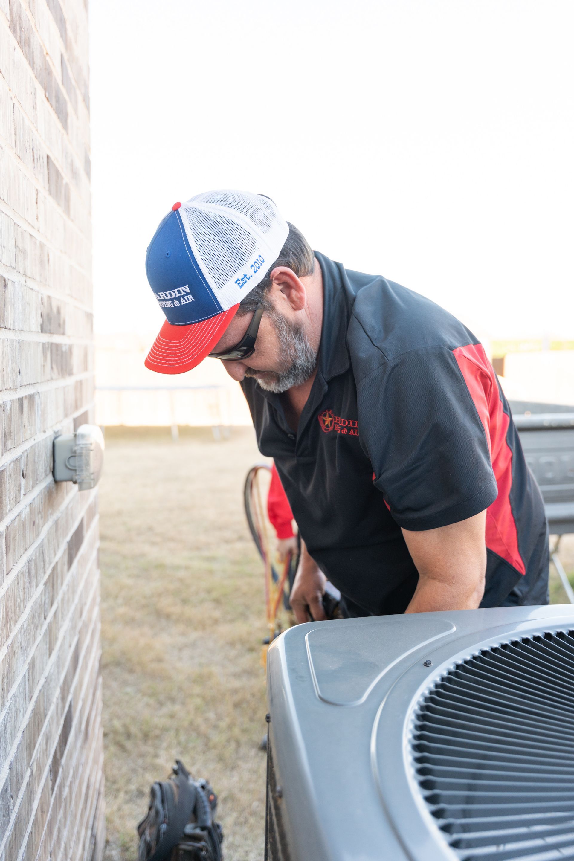 HVAC technician inspecting an outdoor AC unit next to a brick wall, wearing a hat, shirt, and sunglasses.