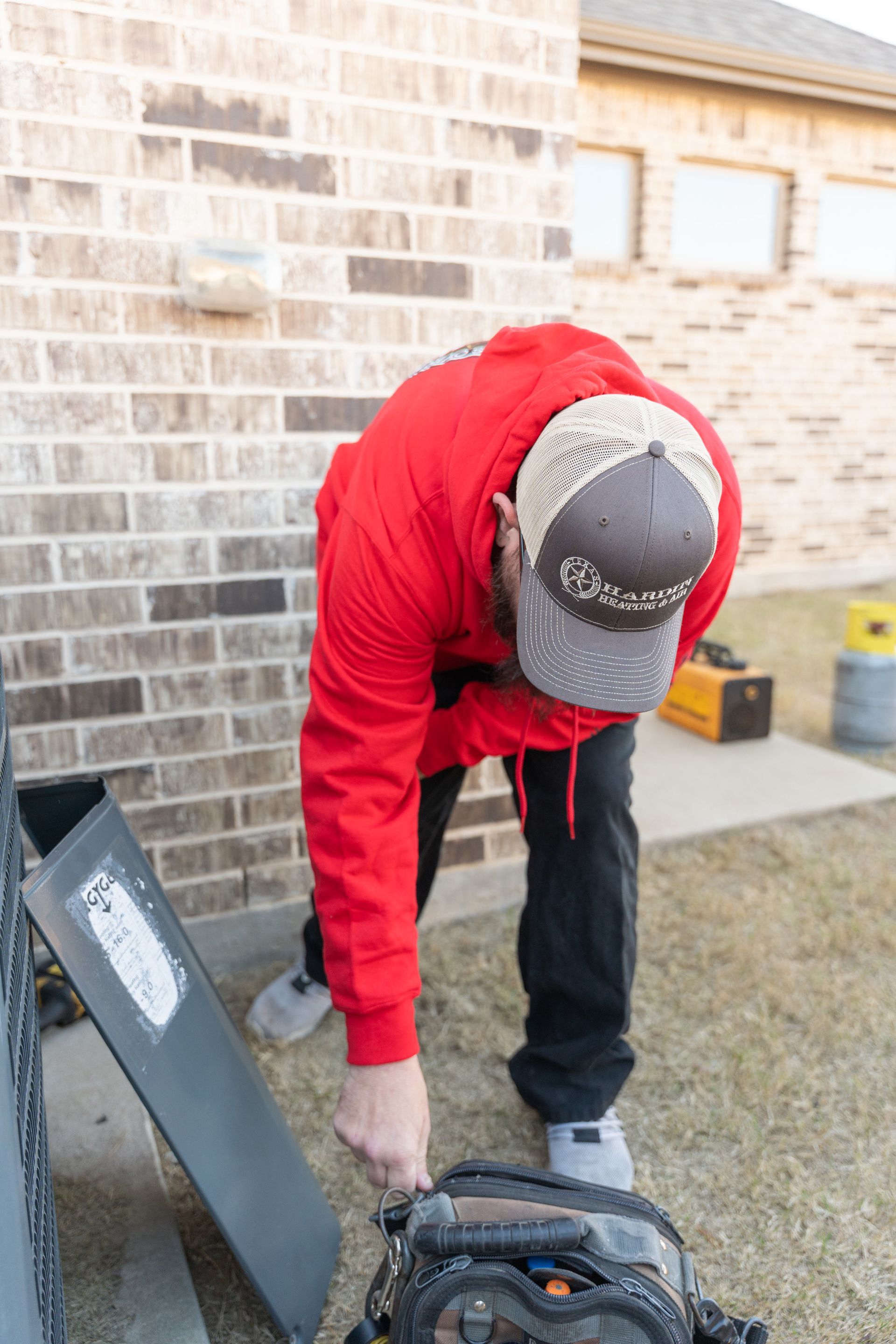 Hardin Heating & Air Technician in red hoodie and baseball cap, working outside near brick wall.
