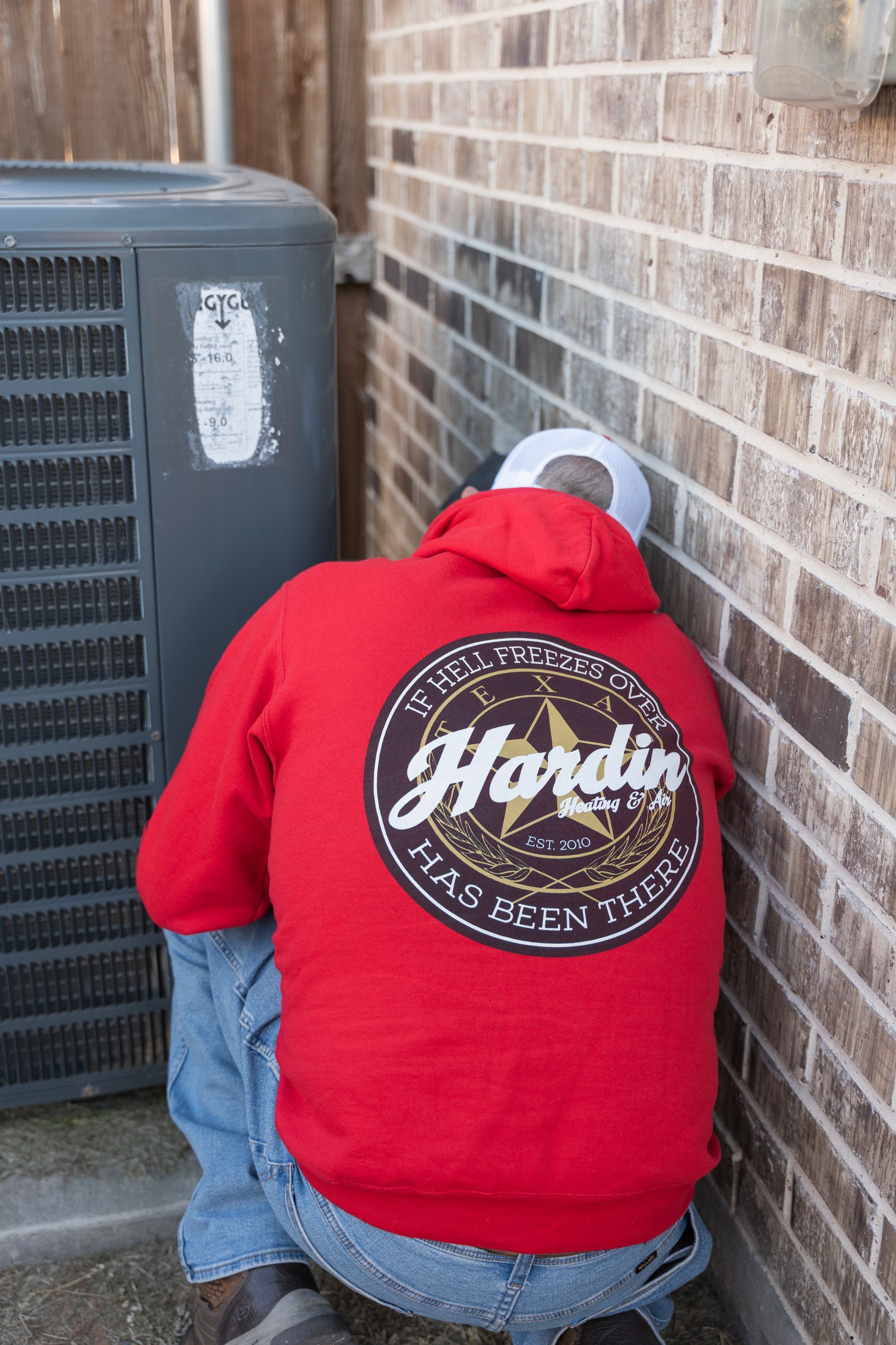 Person in red hoodie inspecting an AC unit against a brick wall.