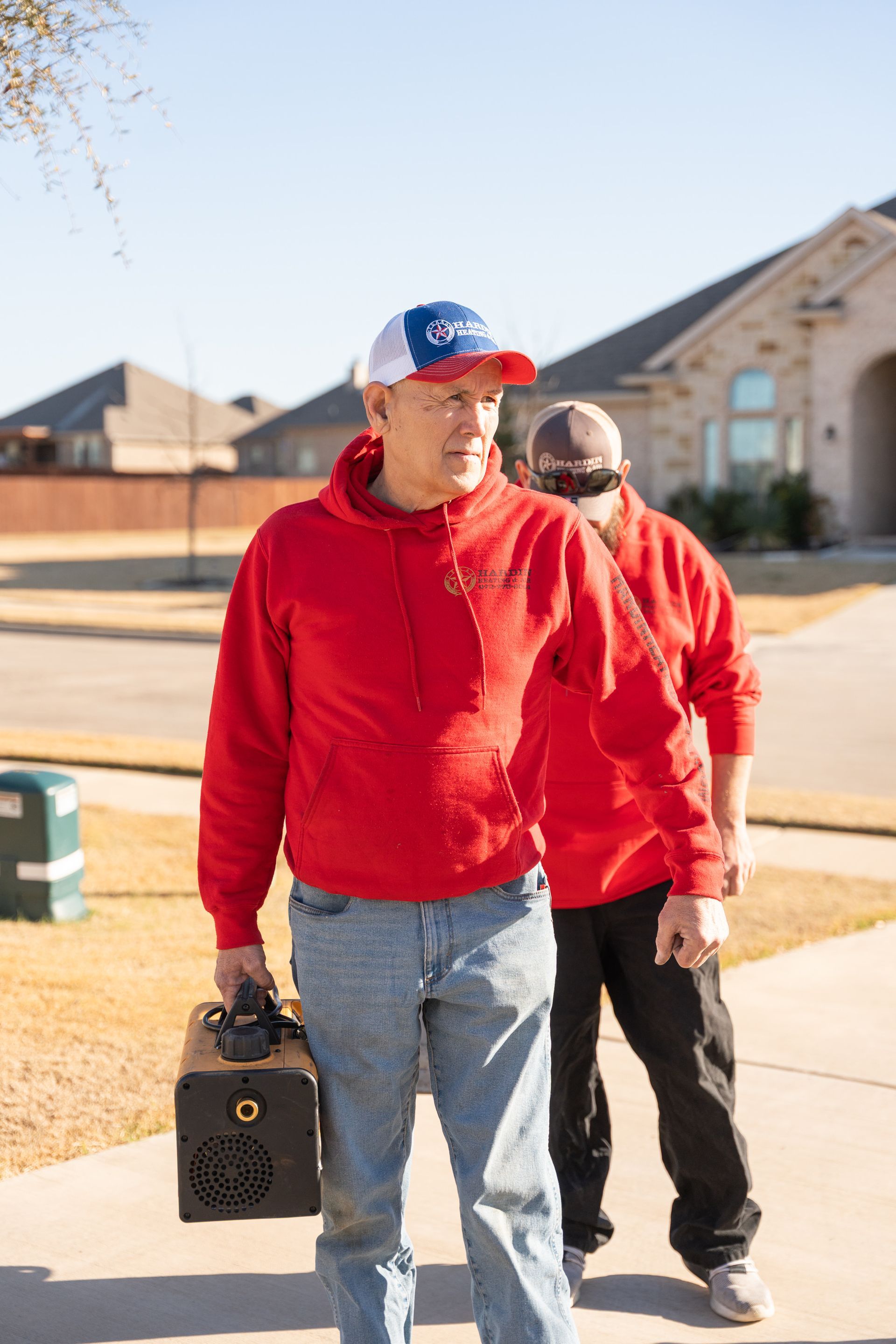 Hardin Heating & Air Technician in red hoodies and jeans on a driveway; one holds a toolbox, the other has a blindfold.