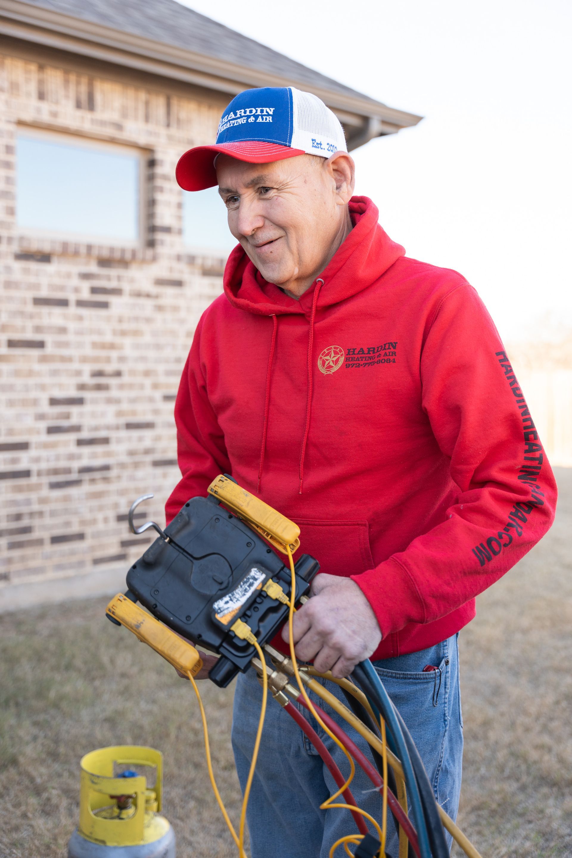 Man in red hoodie and baseball cap holding HVAC tools outside a brick building.