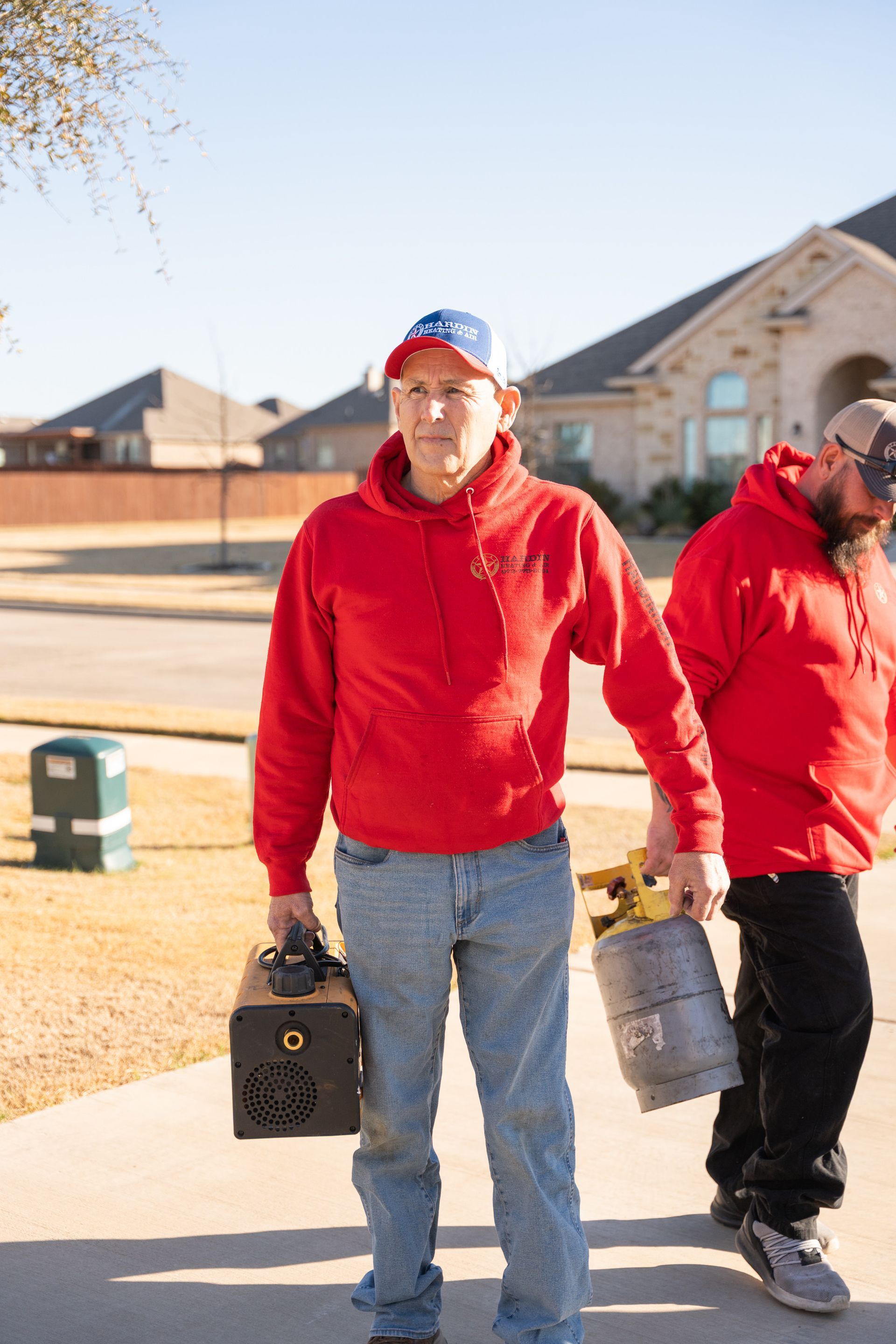 Hardin Heating & Air Technician in red hoodies walking on a sidewalk, one holding a black box, the other a yellow tank.