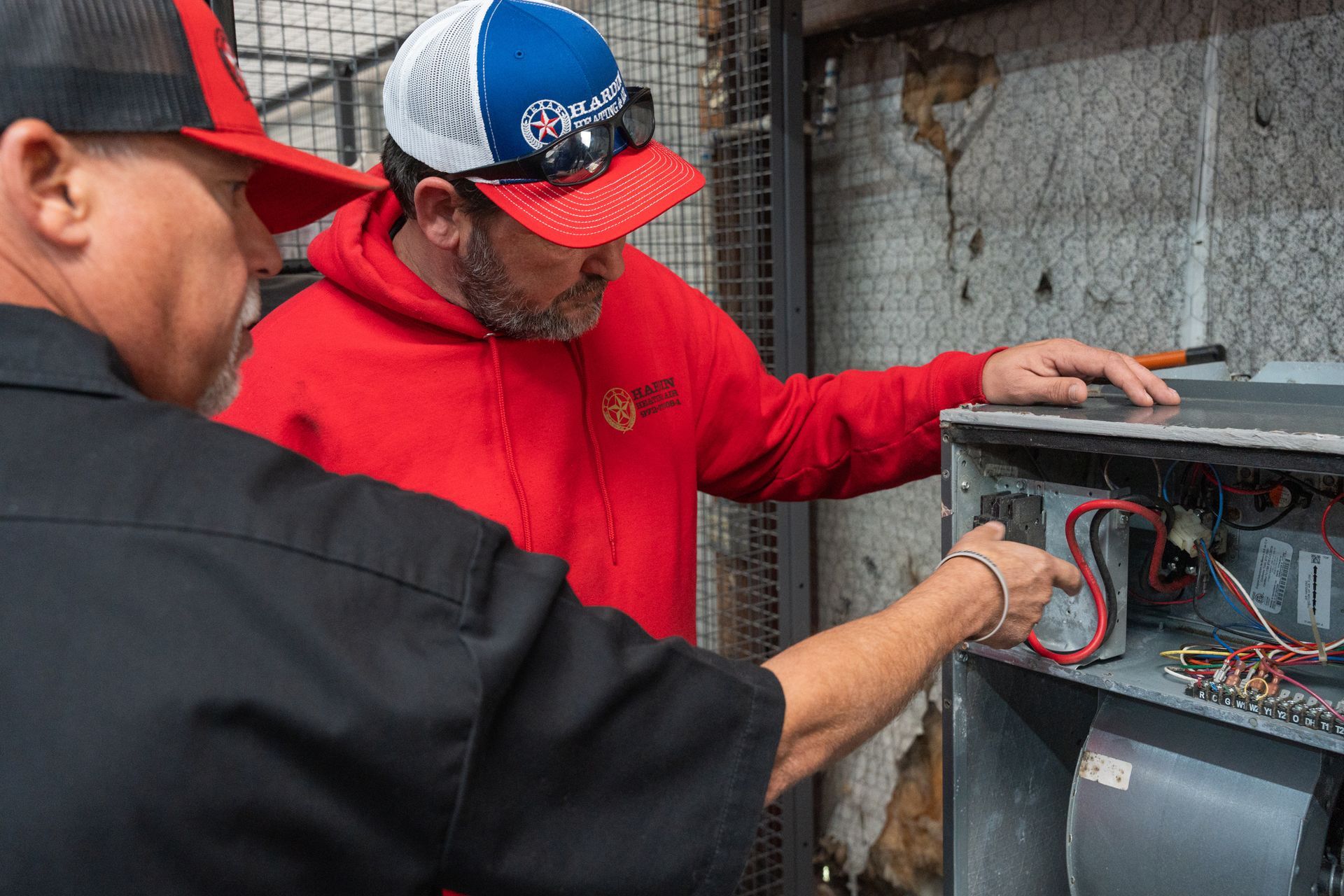 Two Hardin Heating & Air Technician in caps examining electrical components of an HVAC unit.