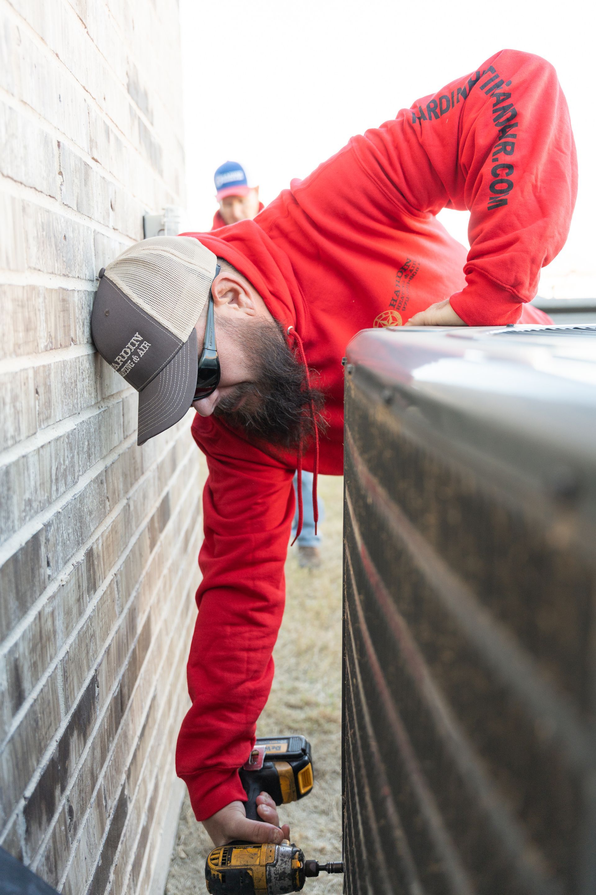 Hardin Heating & Air Technician leans to work on an air conditioner unit between a brick wall, holding a power drill.