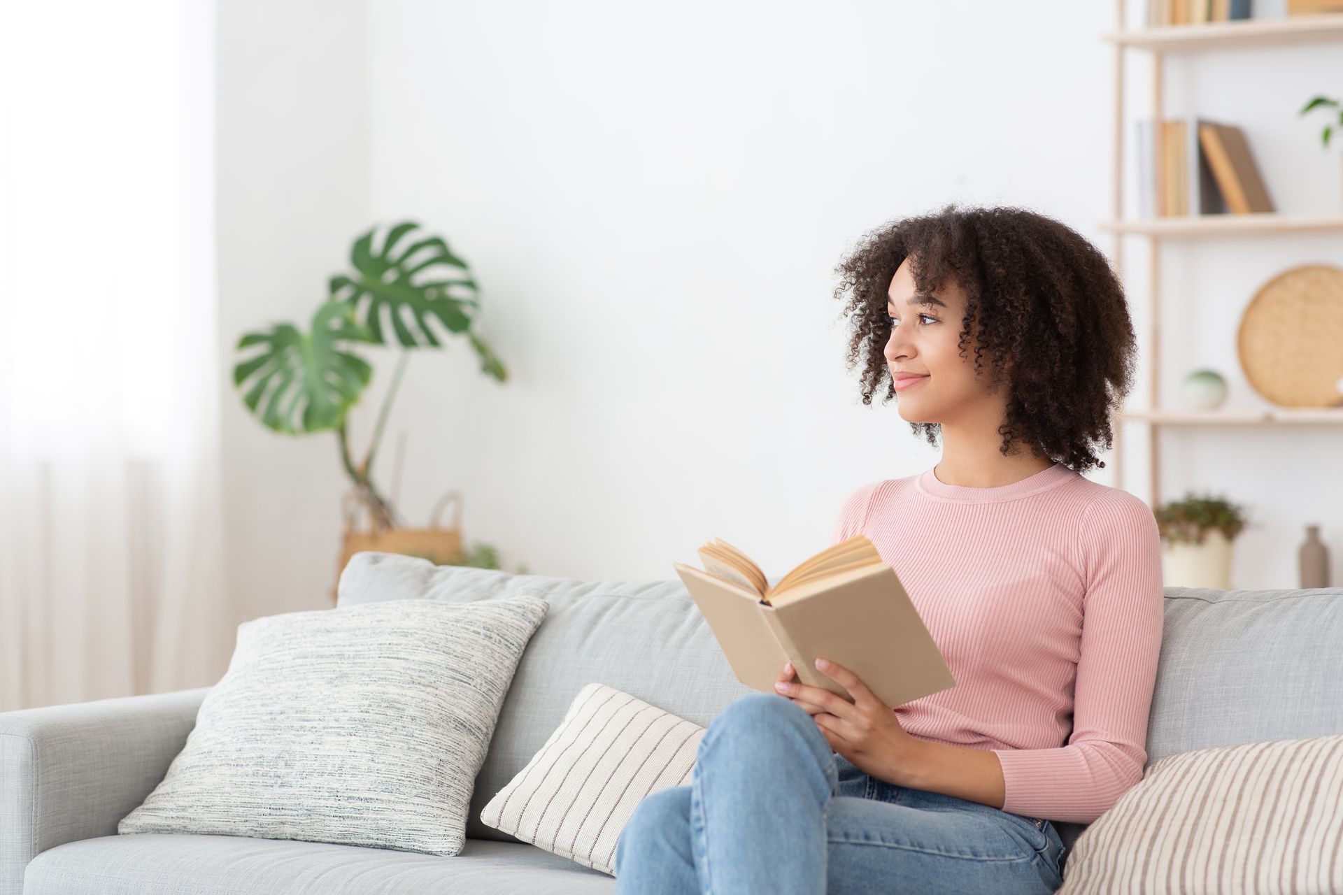 Woman with curly hair reading a book on a gray sofa, looking out a window. Pink shirt, jeans. Bright, modern living room.