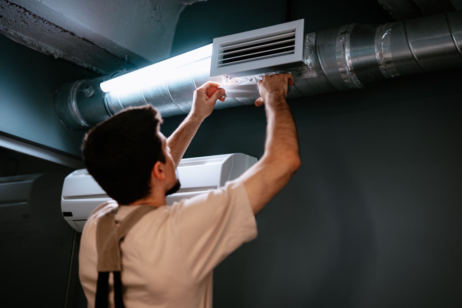 Person adjusting air vent on a duct, with AC unit below and fluorescent light above.