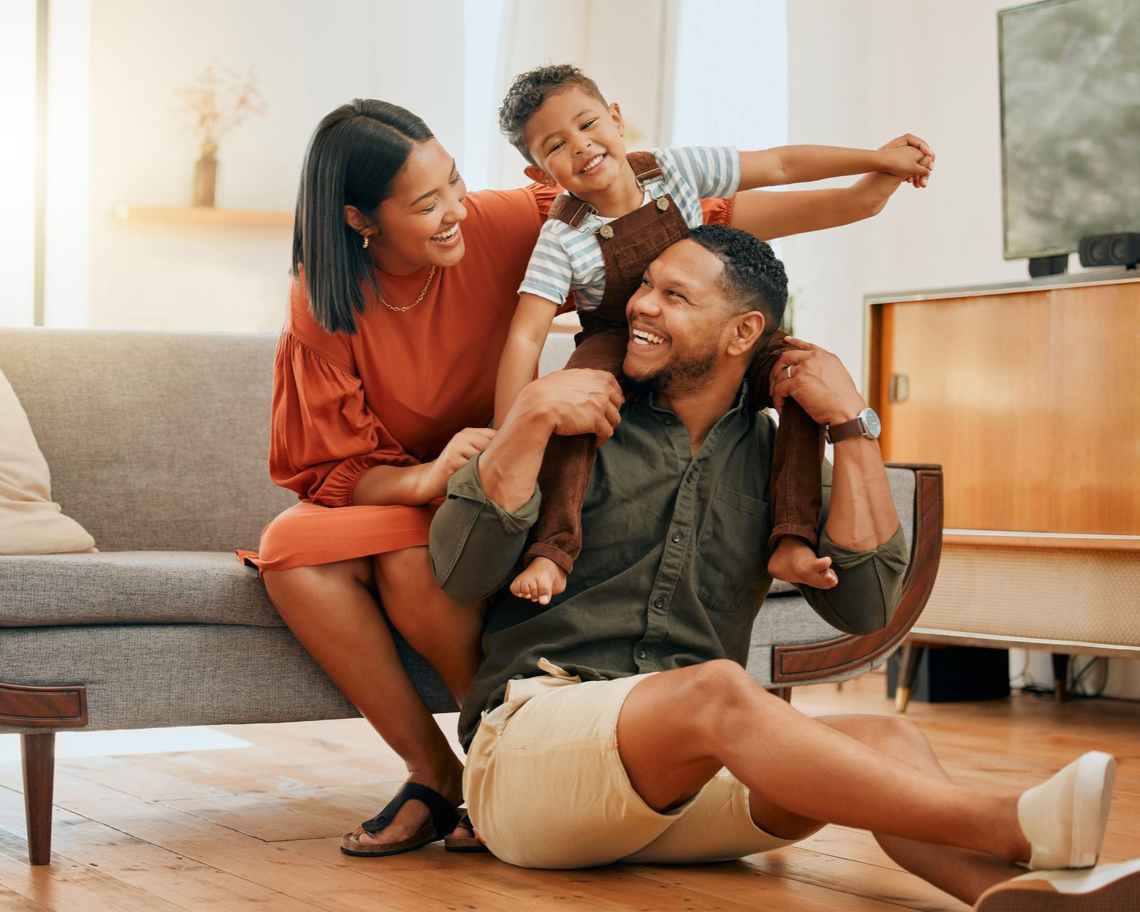 Family smiling together indoors; child on father's shoulders, mother beside them on couch.