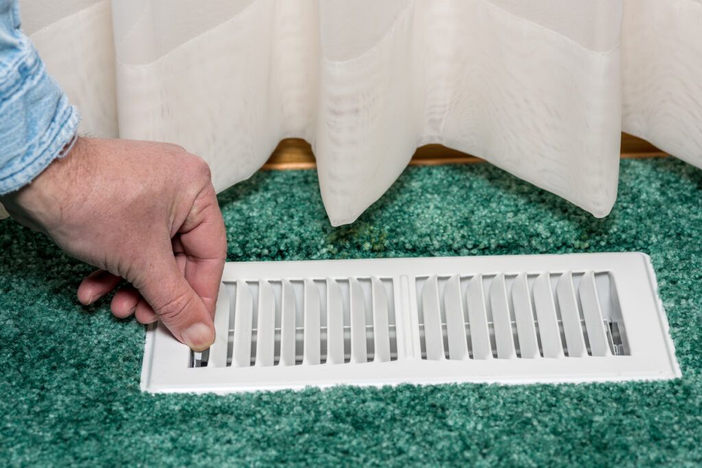 Hand adjusting a white vent cover on a green carpet, near a white curtain.