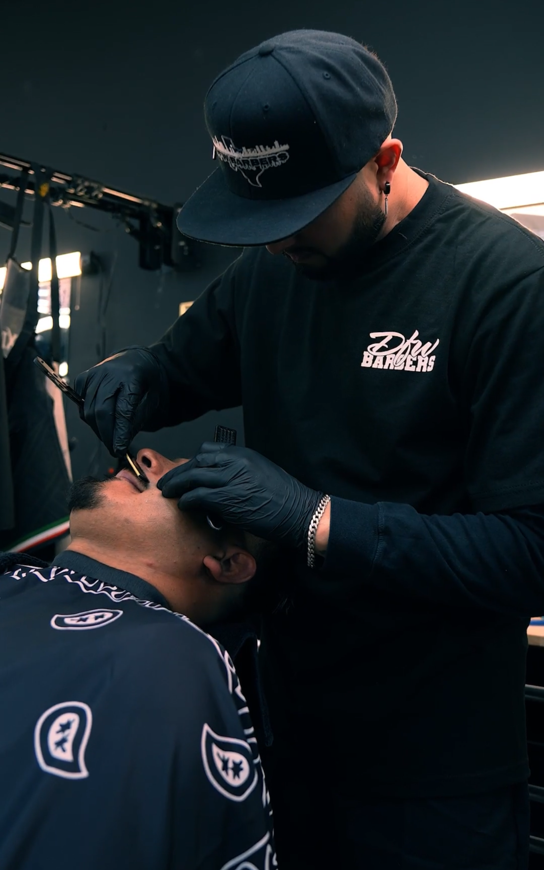 A barber wearing black gloves and a cap uses a straight razor to shave a customer wearing a black paisley cape.