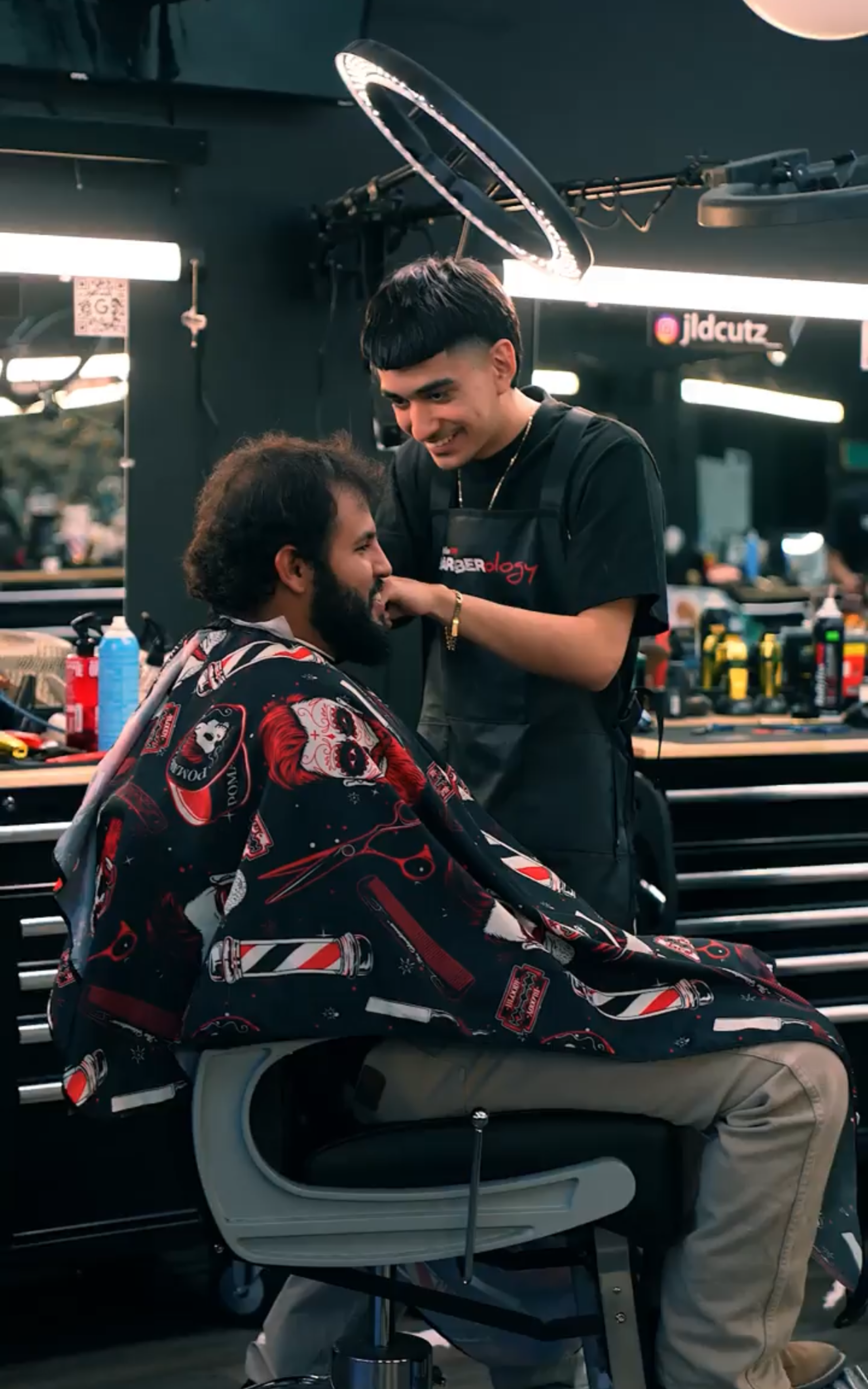 A barber smiles while trimming the hair of a customer sitting in a chair wearing a patterned barber cape in a salon.