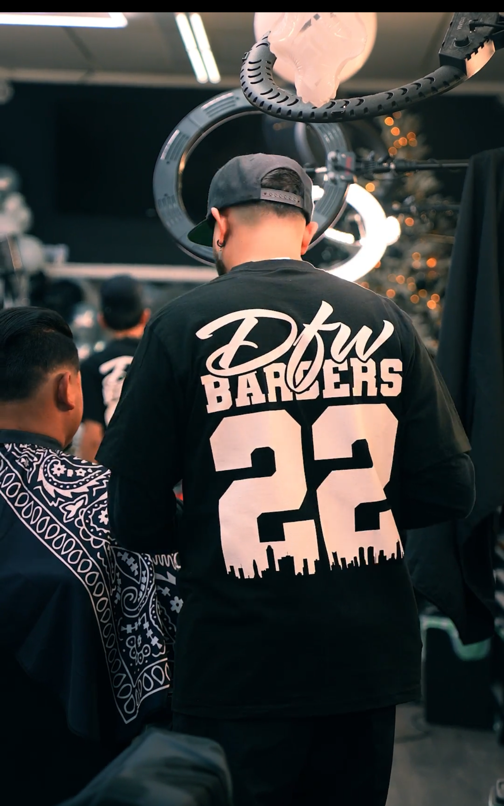 A barber wearing a black long-sleeve shirt with "DFW BARBERS 22" on the back works on a client in a barbershop.