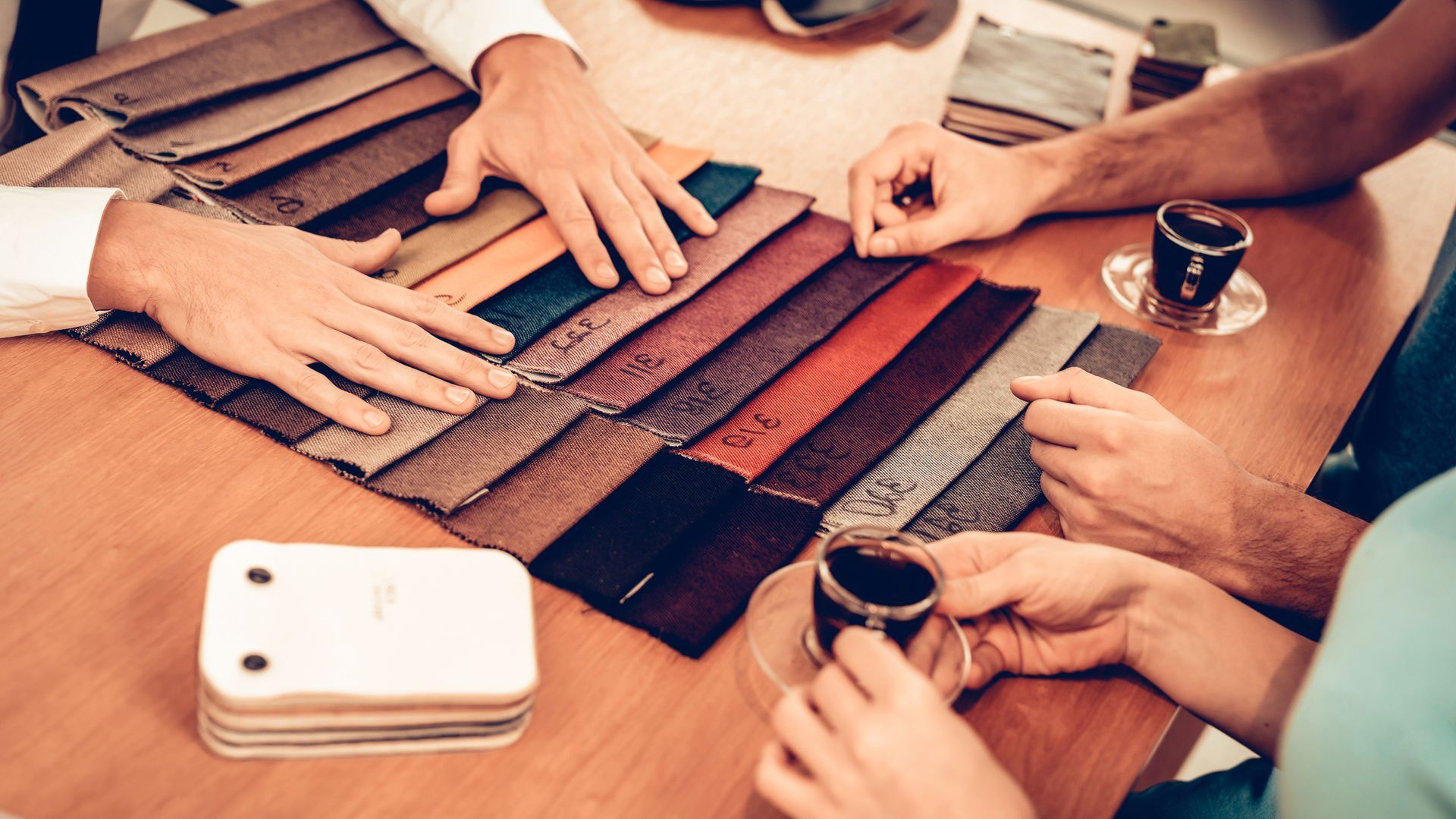 Hands review a spread of fabric swatches on a table while discussing upholstery options over coffee.