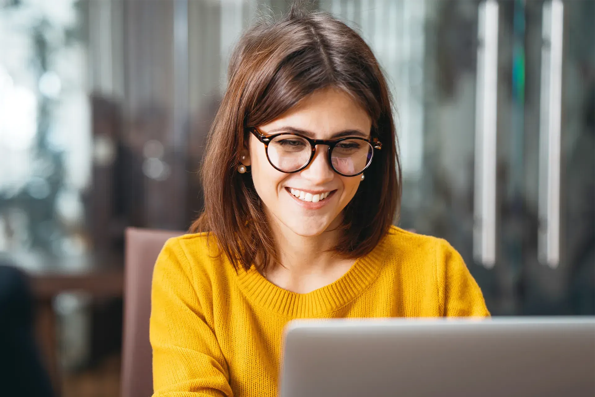 Smiling woman wearing glasses and a mustard yellow jumper, working on a laptop in a modern office se