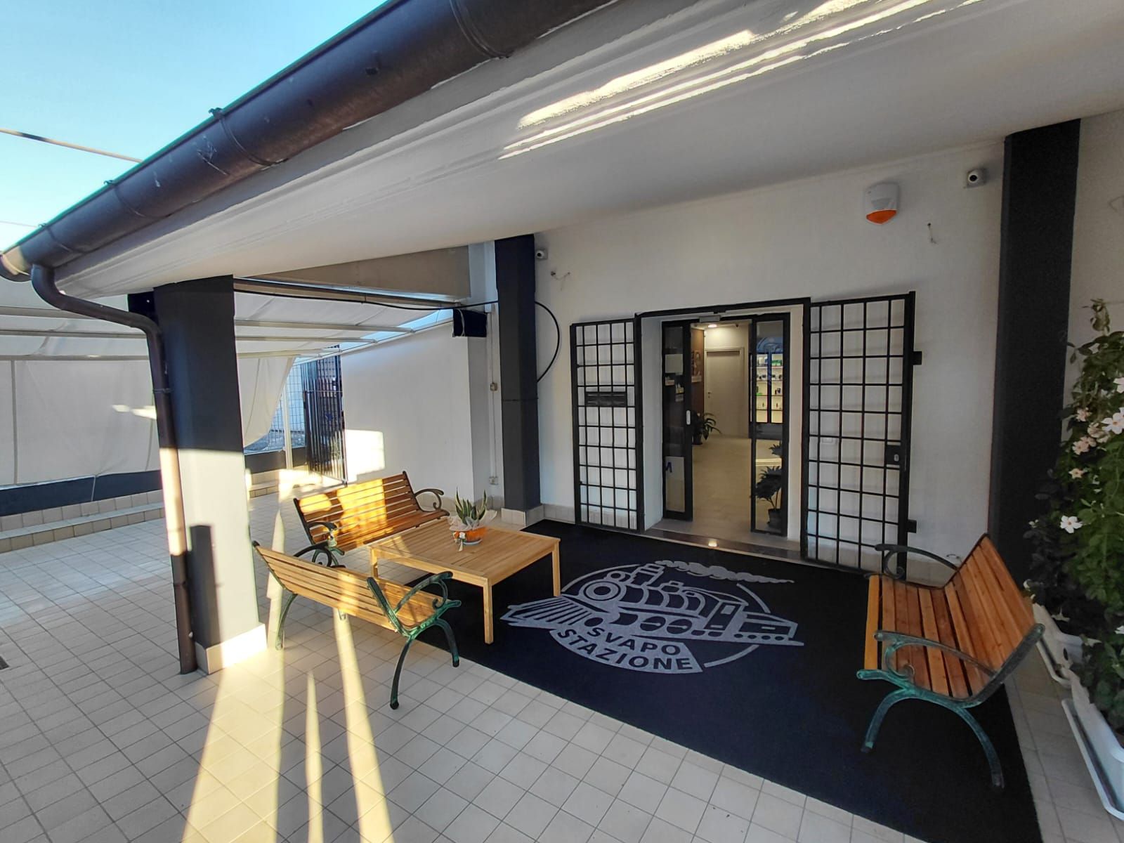 Patio coperto con posti a sedere, che conduce all'ingresso con porte di sicurezza in metallo; zerbino di benvenuto.