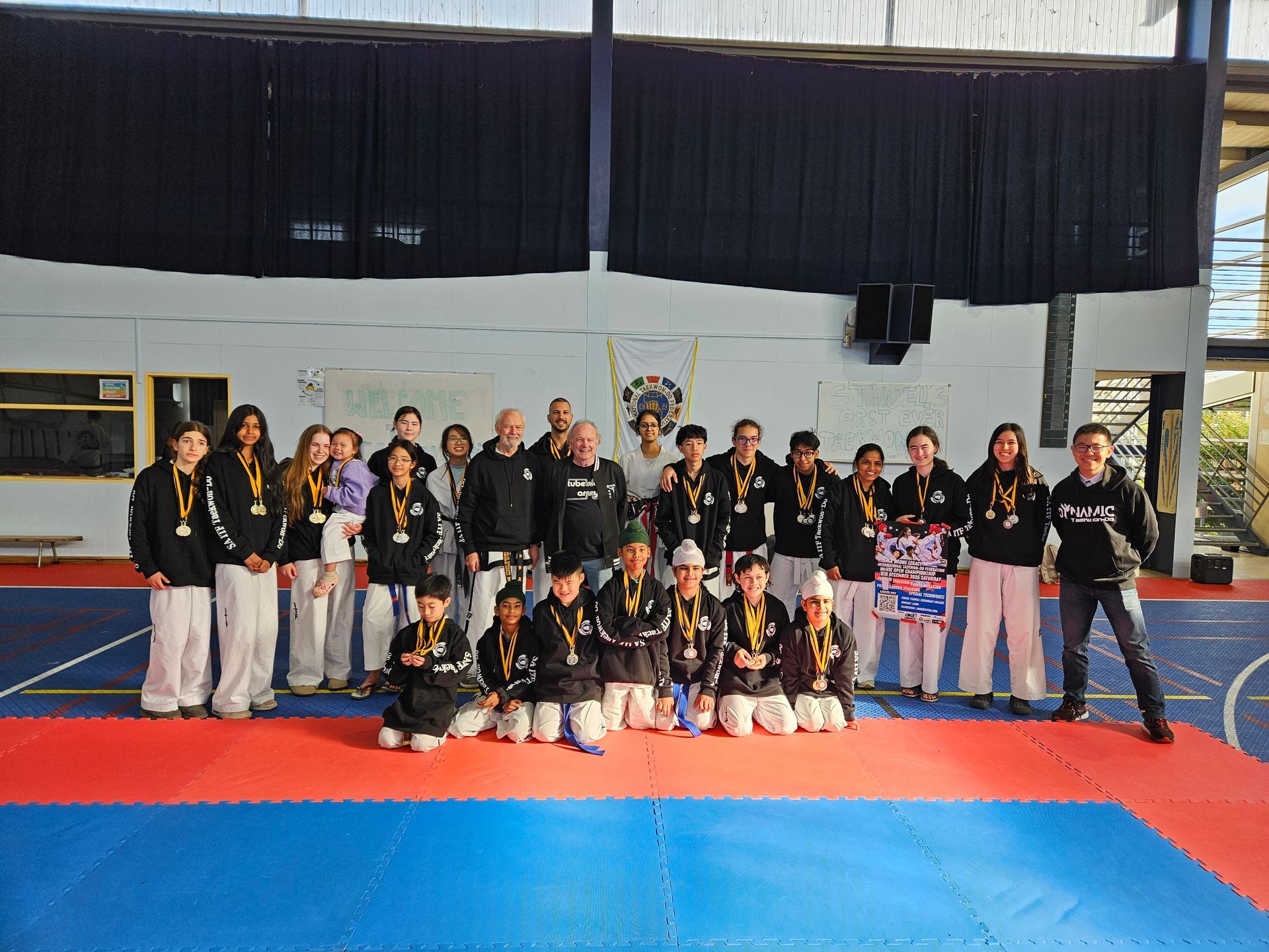 Group posing on blue-and-red mats in a gym, wearing black and white martial arts uniforms