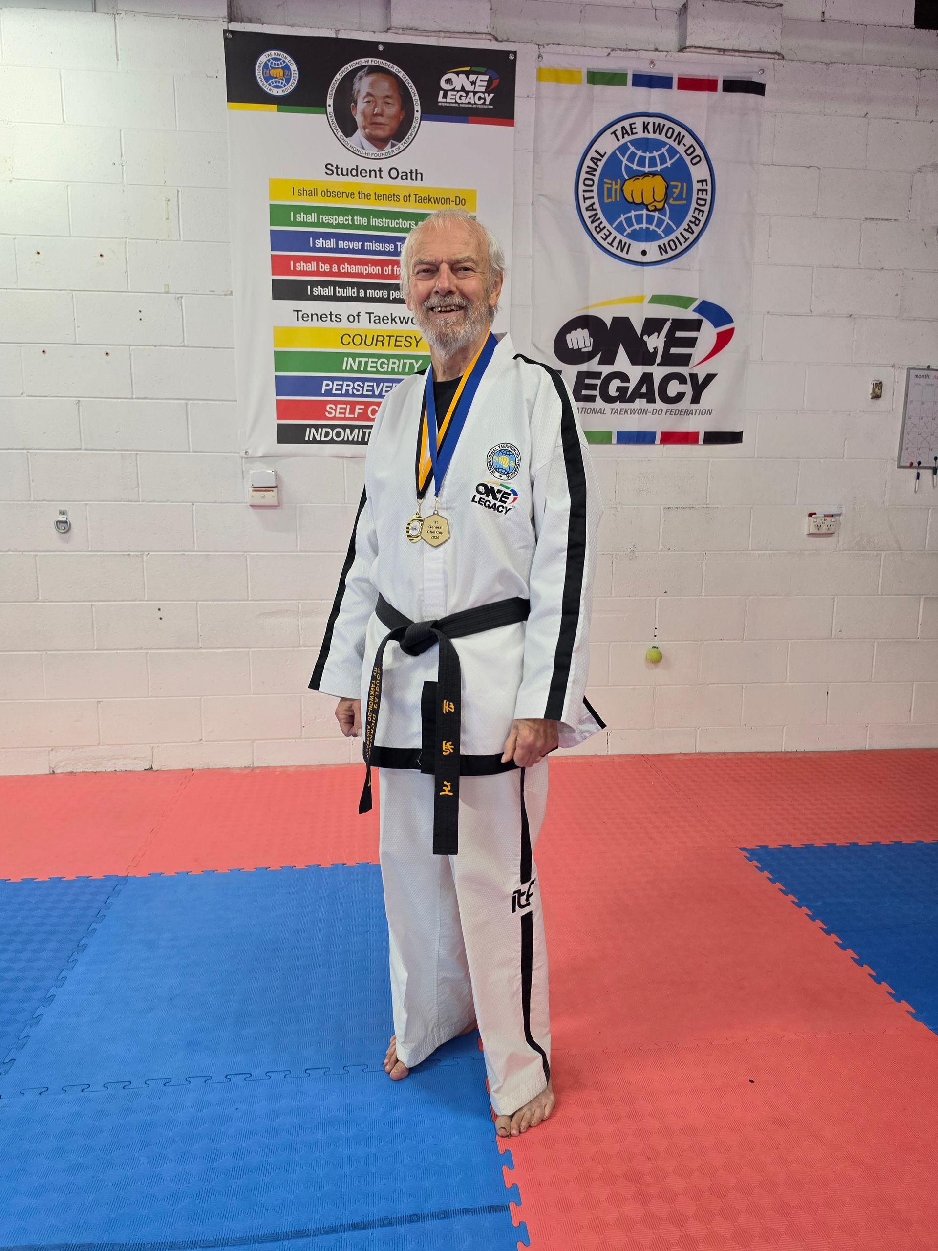Martial artist in white gi and black belt smiling on red-and-blue mats in a dojo.