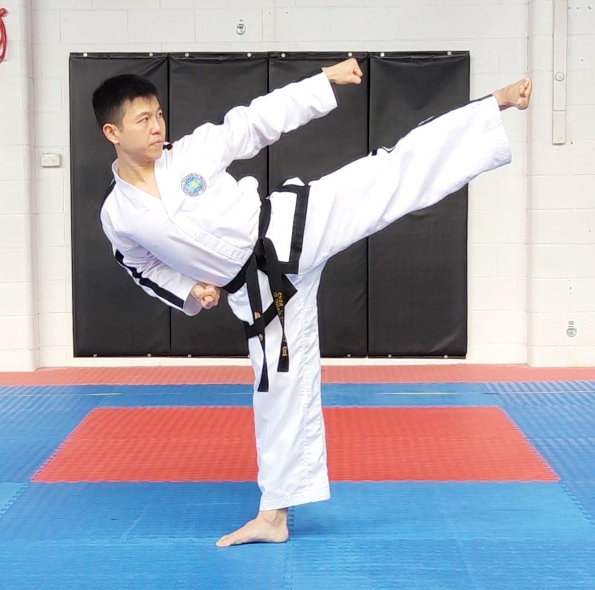 Martial artist in white gi performing a high side kick on blue and red mats indoors