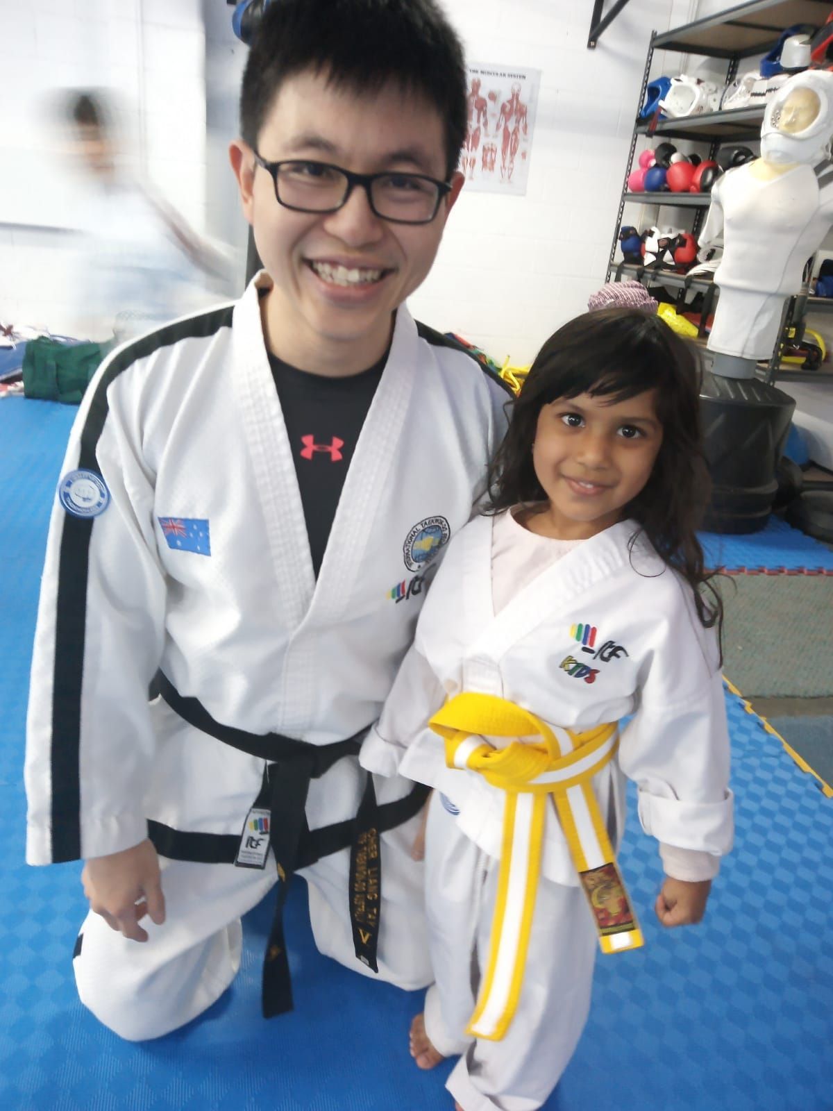 Two people in white martial arts uniforms smiling indoors, one wearing a black belt and the other a yellow belt