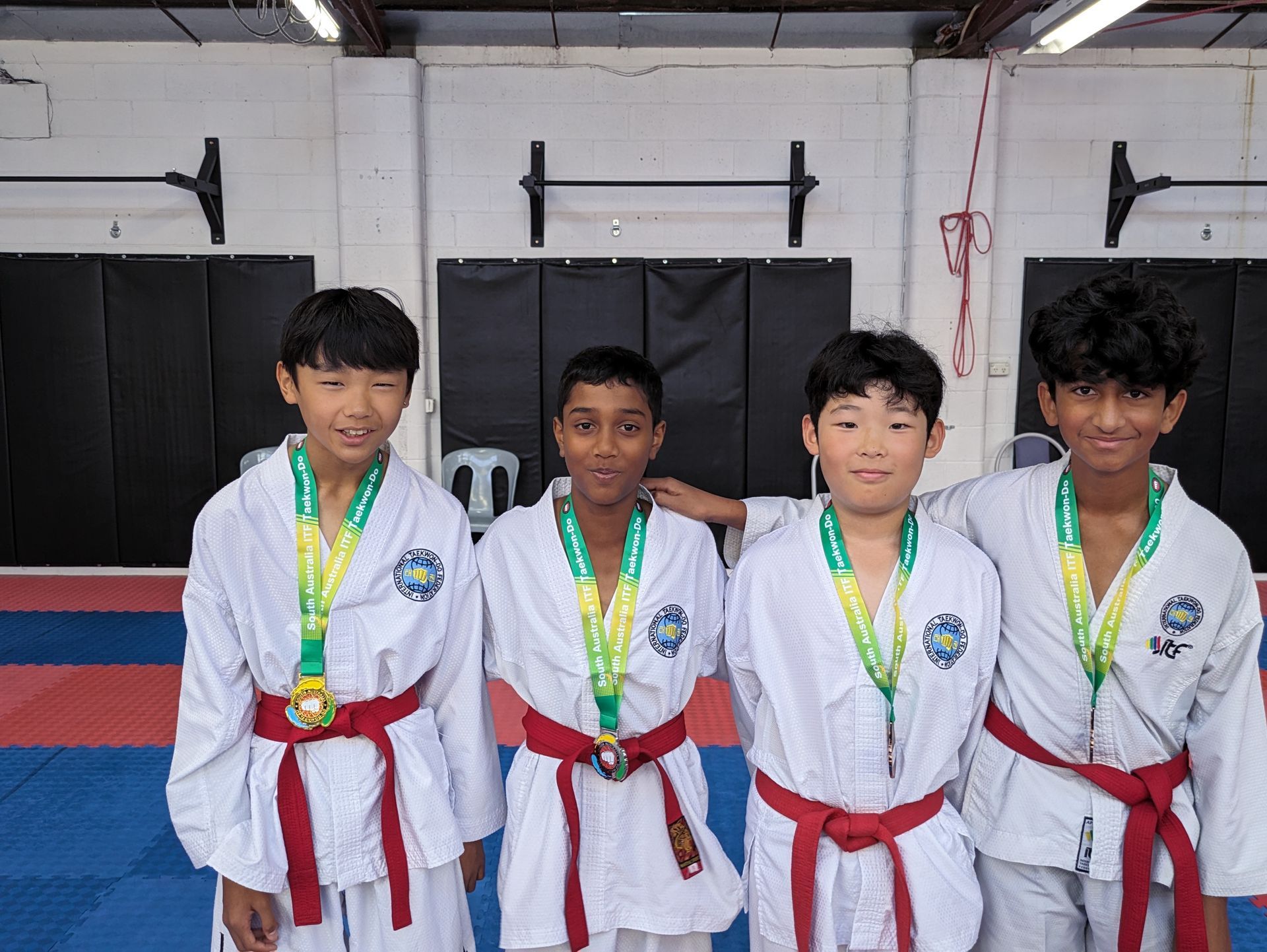 Four martial arts competitors in white gis with red belts and medals standing indoors and smiling