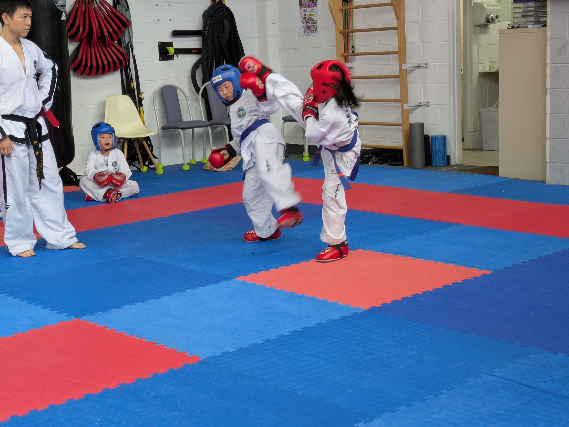 Two children sparring in protective gear on a blue and red martial arts mat, with others watching nearby