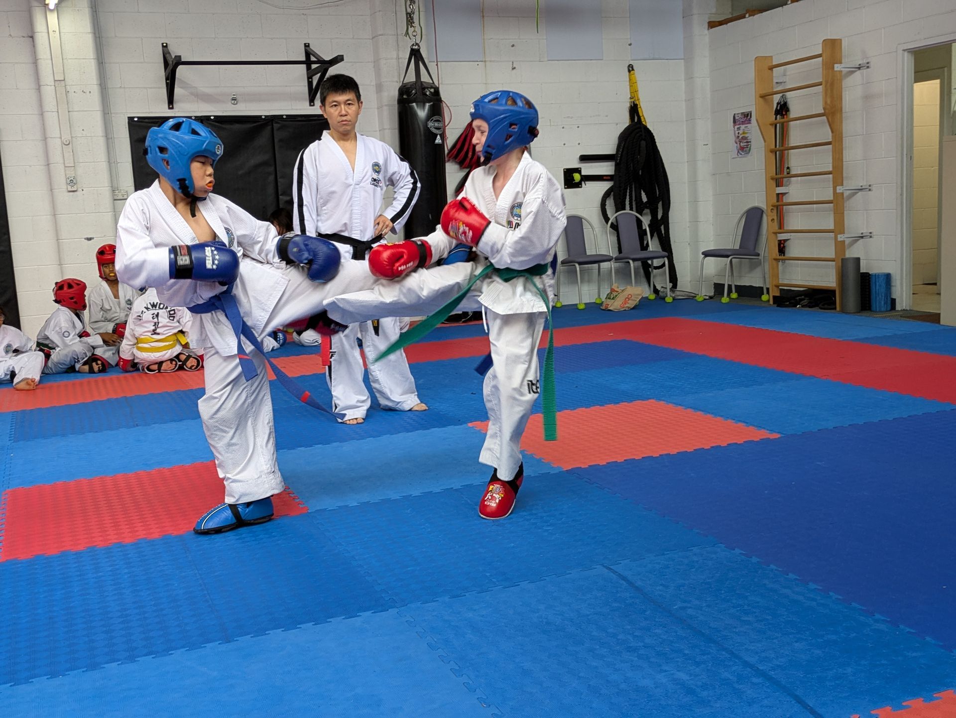 Two martial artists sparring on blue mats while a coach watches in a dojo