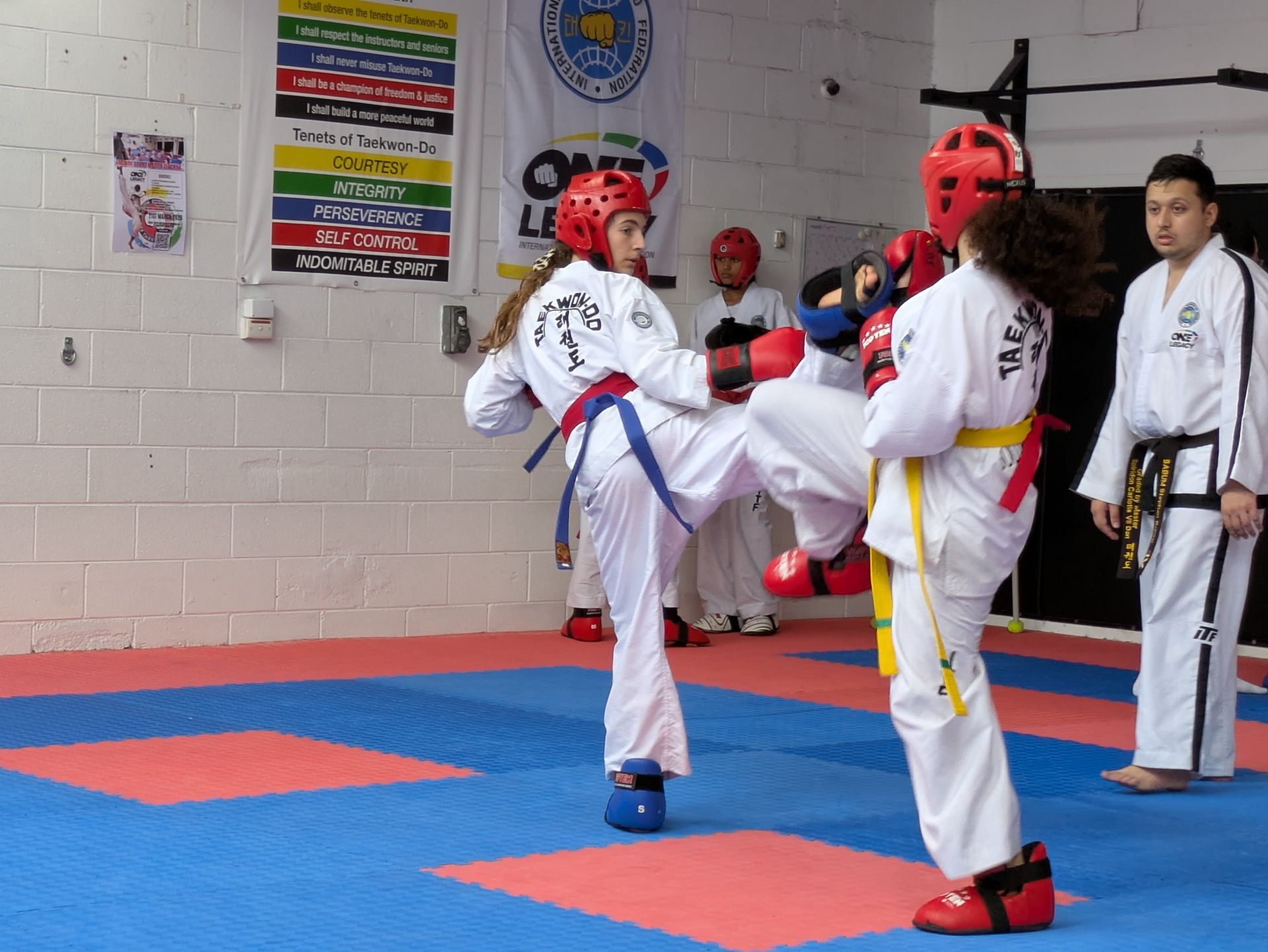 Two taekwondo sparring students in white uniforms and red helmets kick in a gym; instructor watches nearby