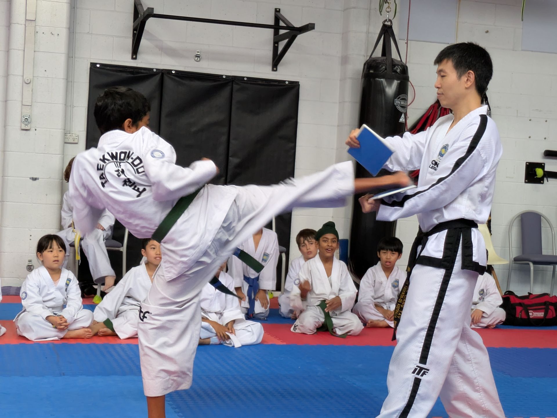 Martial arts student performing a high kick during a belt test while an instructor holds a target board
