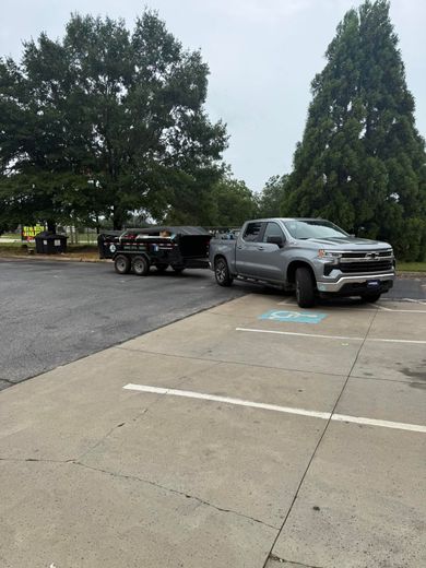 A silver pickup truck towing a black dump trailer parked in a paved parking lot near trees.