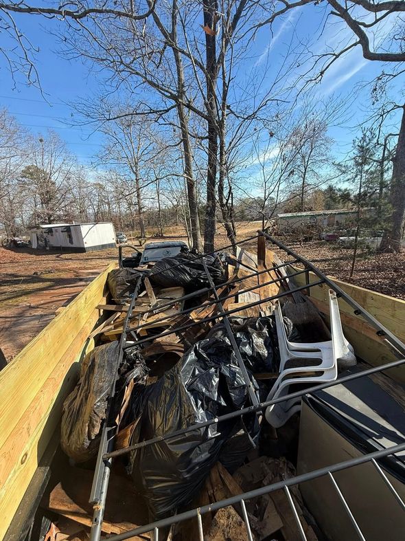 A trailer bed filled with black trash bags, a metal bed frame, and white plastic chairs in a wooded, sunny area.