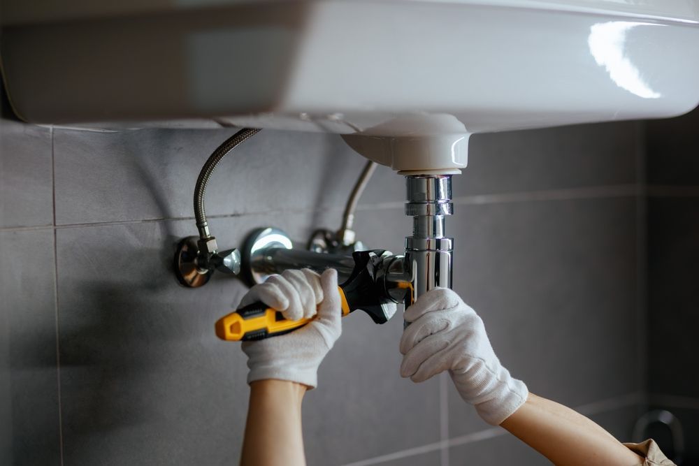 Person in white gloves using a wrench to repair the chrome P-trap under a bathroom sink against a tiled wall.