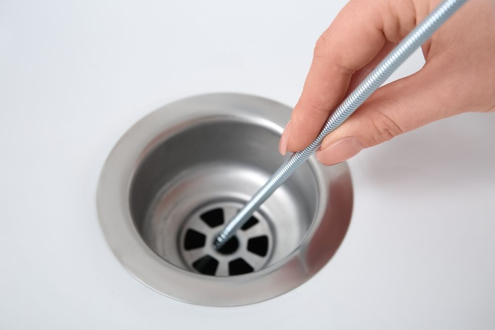A hand holds a metal rod, inserting it into the holes of a silver sink drain stopper in a white sink basin.