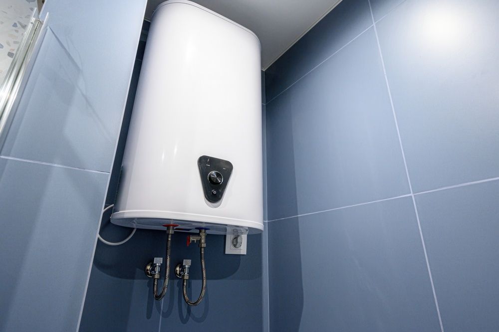 A white electric water heater mounted on a wall with blue tiles, featuring a central control knob and plumbing below.