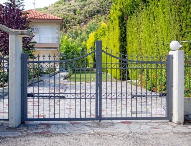 Gray metal gate in front of a house. Tall green hedges in the background. Paved driveway and sidewalk.