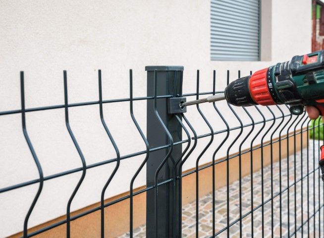 Person using a power drill to attach a section of dark metal fence to a post.