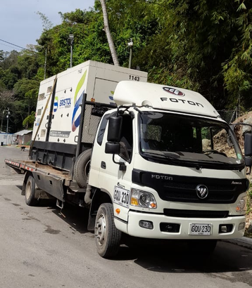 A white Foton flatbed truck carrying a large industrial generator on a road with trees in the background.