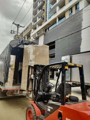A red forklift unloads a large, cardboard-wrapped cargo crate from a truck outside a tall modern apartment building.