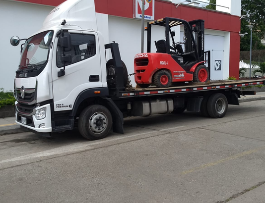 A flatbed truck transporting a red forklift parked on a street in front of a building.