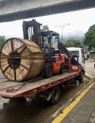 A flatbed tow truck carrying a large wooden wire spool and an orange forklift on a road.