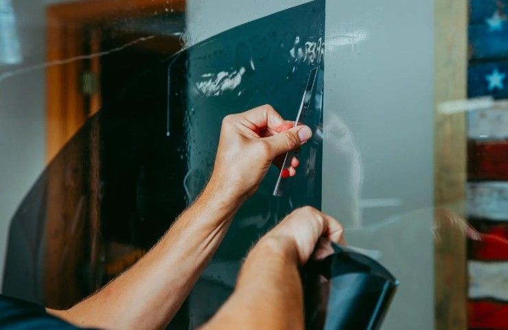 Close-up of hands applying a dark, adhesive window tint film to a glass surface using a squeegee tool.