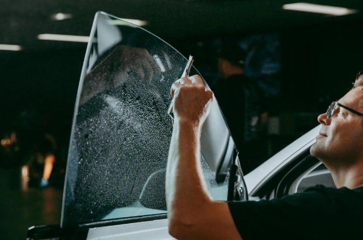 A person uses a squeegee to smooth a wet layer of window tint onto a car side window in a garage.