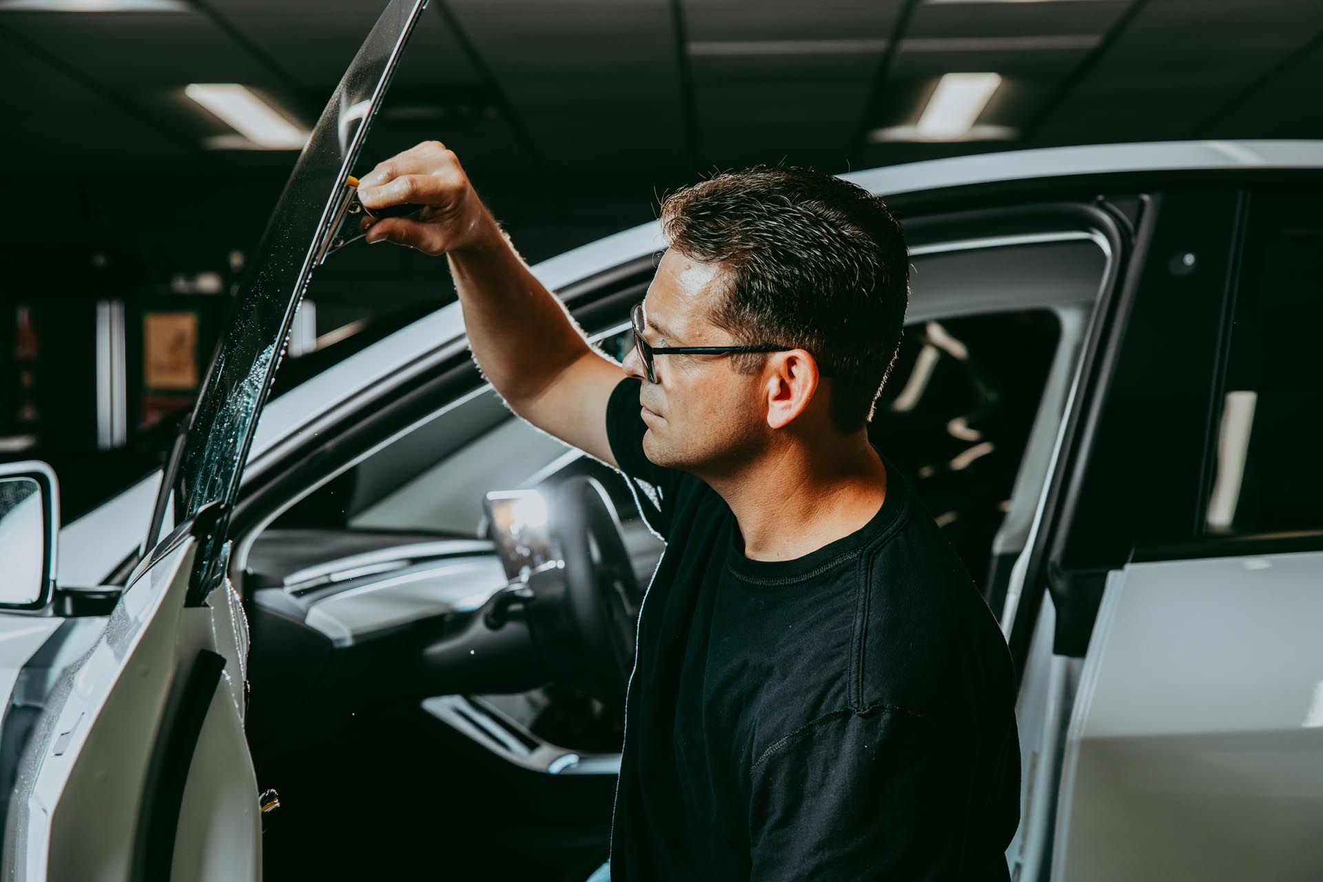 Man inspecting a car windshield in a dim garage, holding the open door.