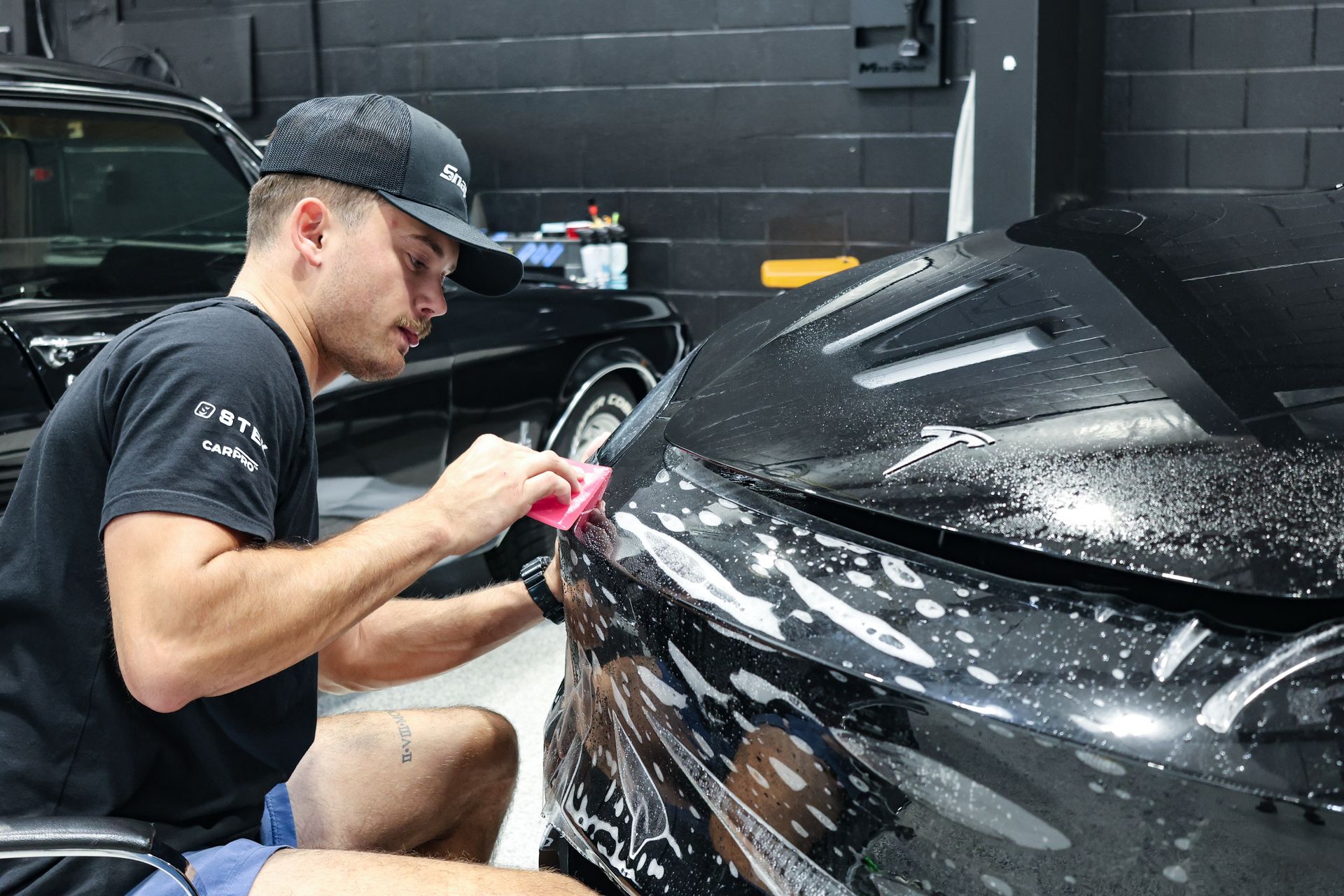 Man applying film to black car's front. He's kneeling in a garage, holding a tool, working on paint protection.